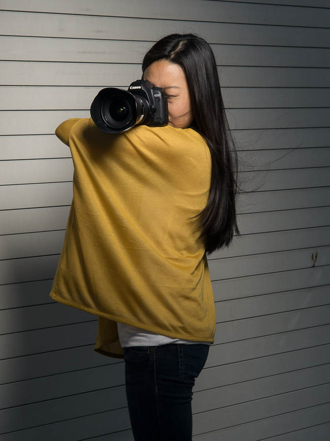 Photographer demonstrating the elbow shelf technique by gripping the right shoulder with the left hand and resting the camera on the left arm
